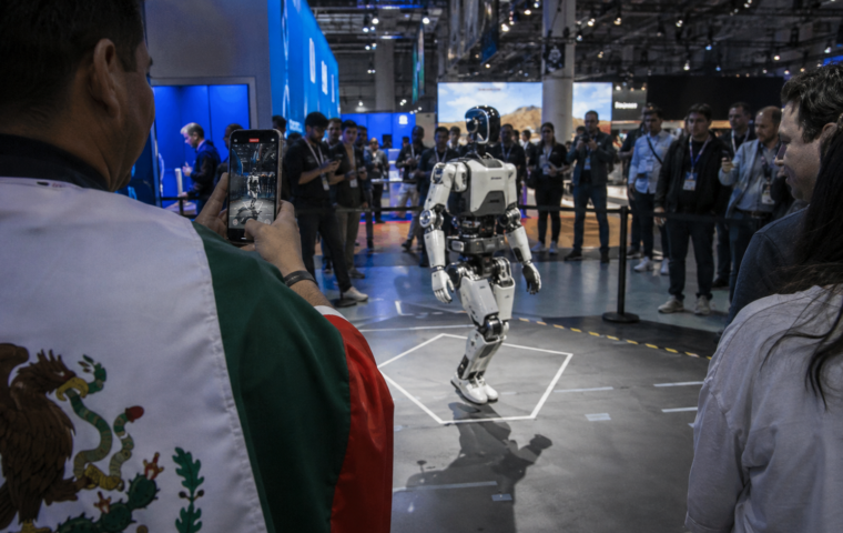 Visitante graba a un robot humanoide en una feria tecnológica, con asistentes alrededor y una persona con referencia mexicana en su ropa.
